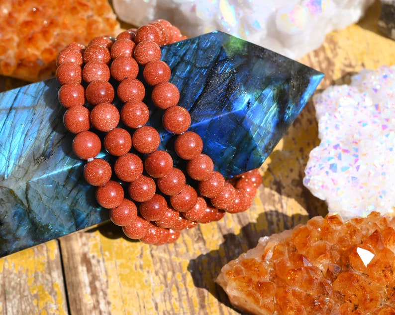 Puede incluir: Primer plano de un brazalete de cuentas de varias hebras hecho de cuentas de piedra arenisca rojo-marr&oacute;n natural. El brazalete est&aacute; sobre un cristal de labradorita azul, rodeado de otros cristales, incluyendo un c&uacute;mulo de cuarzo blanco y un c&uacute;mulo de citrino amarillo.