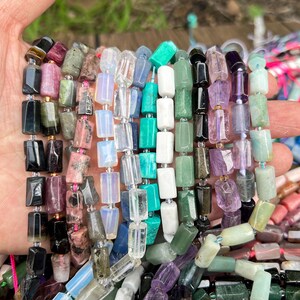 May include: A hand holding multiple strands of gemstone beads in various colors. The beads are rectangular and include shades of black, pink, green, white, and purple. The beads are strung together, ready for jewelry making.
