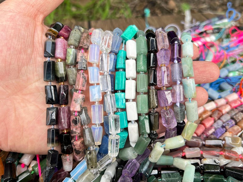 May include: A hand holding multiple strands of gemstone beads in various colors. The beads are rectangular and include shades of black, pink, green, white, and purple. The beads are strung together, ready for jewelry making.
