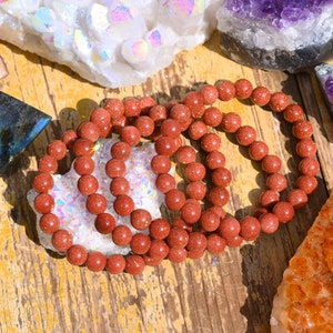 Puede incluir: Un conjunto de cuatro pulseras de piedras preciosas de color rojo-marr&oacute;n hechas de cuentas redondas. Las pulseras est&aacute;n apiladas una encima de la otra y est&aacute;n sobre una superficie de madera.