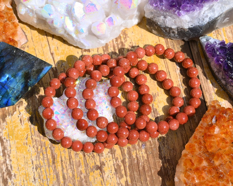 Puede incluir: Un conjunto de cuatro pulseras de piedras preciosas de color rojo-marr&oacute;n hechas de cuentas redondas. Las pulseras est&aacute;n apiladas una encima de la otra y est&aacute;n sobre una superficie de madera.
