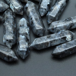 Puede incluir: Un grupo de puntas de cristal de labradorita negra, también conocidas como piedra lunar negra, con un efecto brillante e iridiscente. Las puntas están dispuestas en un patrón aleatorio sobre una superficie gris oscuro.