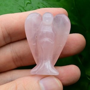 Puede incluir: Peque&ntilde;a figura de &aacute;ngel de cuarzo rosa transl&uacute;cido. El &aacute;ngel tiene alas extendidas y una t&uacute;nica fluida. La piedra es de color rosa suave y los detalles del &aacute;ngel est&aacute;n bien definidos. El fondo es verde borroso.