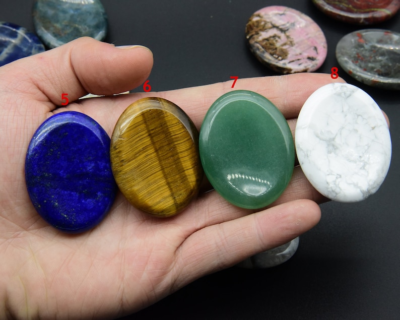 May include: Five smooth, polished oval-shaped stones in various colors. The stones are blue, yellow, green, white, and a mix of brown and pink. The stones are arranged in a row on a hand.