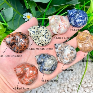 May include: A collection of ten sleeping cat figurines carved from different types of stones, including red obsidian, dalmatian stone, red line, sodalite, branzite, sea quartz, and quartz. Each figurine is unique in color and pattern.