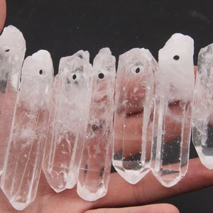 May include: A group of eight clear quartz crystal points with drilled holes for hanging. The crystals are approximately 2 inches long and have a natural, raw finish.