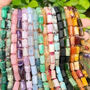 May include: Strands of gemstone beads in various colors, including green, pink, purple, and orange. The beads are rectangular and strung together, showcasing a variety of natural stone textures and hues. The beads are held in a hand.