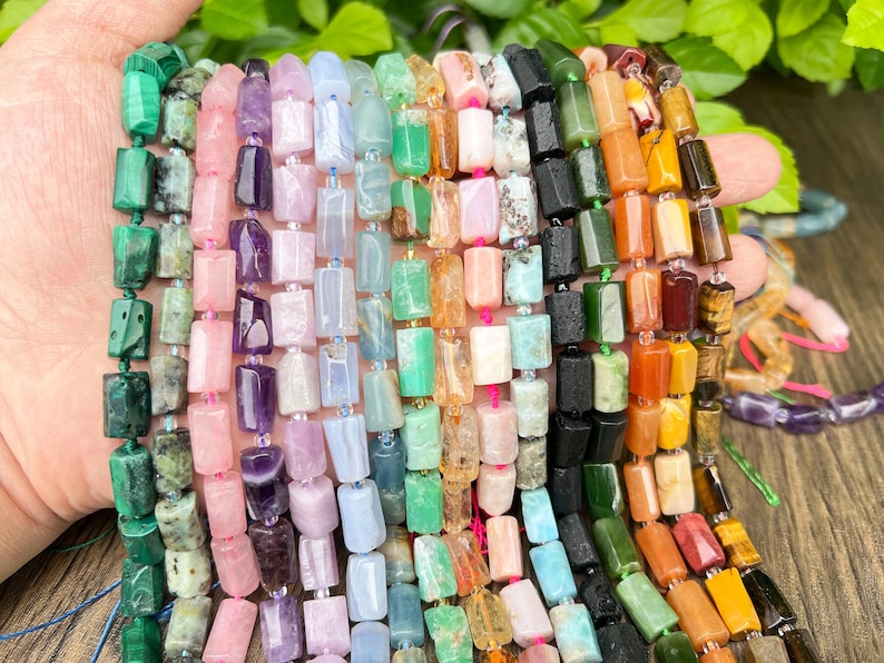 May include: Strands of gemstone beads in various colors, including green, pink, purple, and orange. The beads are rectangular and strung together, showcasing a variety of natural stone textures and hues. The beads are held in a hand.