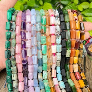 May include: A hand holding strands of colorful gemstone beads. The beads are rectangular and come in various colors, including green, pink, purple, blue, orange, and brown. The beads are strung together, creating a vibrant display of natural stones.