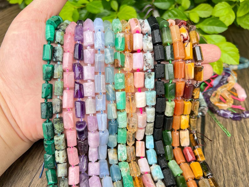 May include: A hand holding strands of colorful gemstone beads. The beads are rectangular and come in various colors, including green, pink, purple, blue, orange, and brown. The beads are strung together, creating a vibrant display of natural stones.