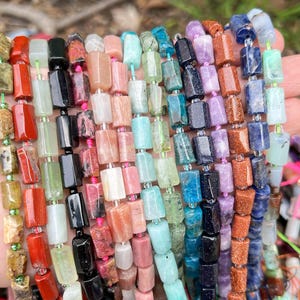 May include: A collection of colorful gemstone bead strands. The beads are rectangular and strung together, showcasing a variety of colors including red, green, black, pink, blue, and purple. The beads are held in a hand, with the background slightly blurred.