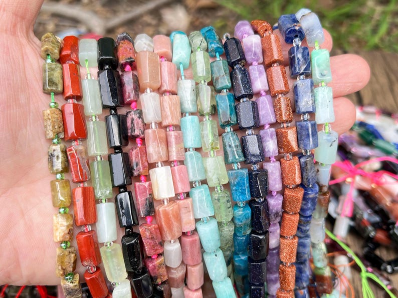May include: A collection of colorful gemstone bead strands. The beads are rectangular and strung together, showcasing a variety of colors including red, green, black, pink, blue, and purple. The beads are held in a hand, with the background slightly blurred.