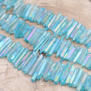 May include: A collection of iridescent blue quartz crystal points, also known as angel aura quartz, arranged in rows on a wooden surface. The crystals are long and slender, with a shimmering, rainbow-like effect.
