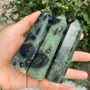 May include: Three green and black patterned crystal points, also known as green moss agate, are held in a hand. The points are smooth and polished.