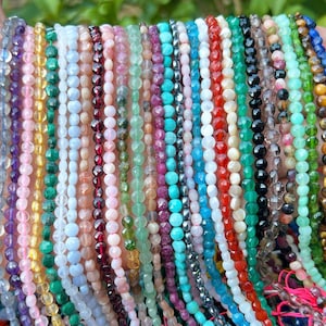 May include: A hand holds a collection of gemstone bead strands in various colors. The strands include amethyst, pink tourmaline, malachite, turquoise, and more. Each strand is made of small, round beads.