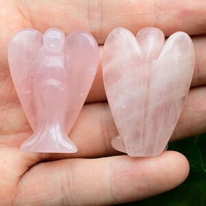 Puede incluir: Dos figuras de &aacute;ngeles de cuarzo rosa. Los &aacute;ngeles est&aacute;n tallados en una piedra rosa p&aacute;lido, con alas detalladas y una t&uacute;nica fluida. Las figuras se sostienen en una mano, mostrando las variaciones naturales de color y la translucidez de la piedra.