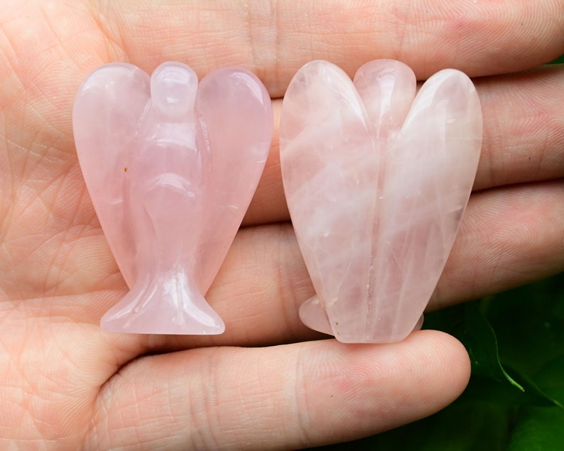 Puede incluir: Dos figuras de &aacute;ngeles de cuarzo rosa. Los &aacute;ngeles est&aacute;n tallados en una piedra rosa p&aacute;lido, con alas detalladas y una t&uacute;nica fluida. Las figuras se sostienen en una mano, mostrando las variaciones naturales de color y la translucidez de la piedra.