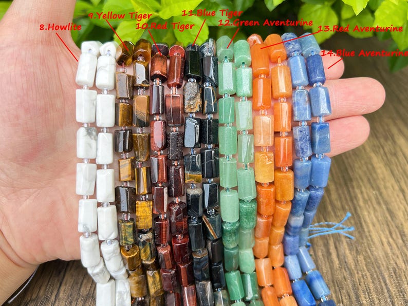 May include: A hand holds multiple strands of rectangular gemstone beads. The beads are in various colors, including white, yellow, red, green, orange, and blue. The gemstones are labeled as Howlite, Yellow Tiger, Red Tiger, Blue Tiger, Green Aventurine, Red Aventurine, and Blue Aventurine.