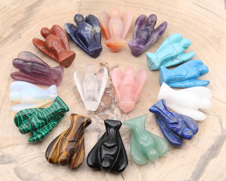 May include: A collection of 14 angel figurines carved from various gemstones, including amethyst, rose quartz, clear quartz, turquoise, and malachite. The figurines are arranged in a circle on a wooden surface.