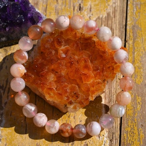 May include: A pink and orange agate gemstone bracelet with a natural, polished finish. The bracelet is made of round beads and is displayed on a wooden surface.