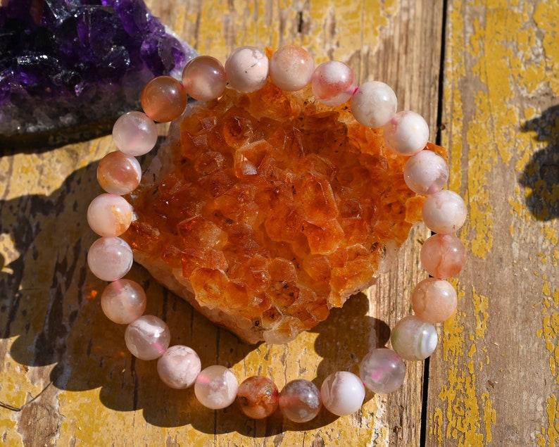 May include: A pink and orange agate gemstone bracelet with a natural, polished finish. The bracelet is made of round beads and is displayed on a wooden surface.