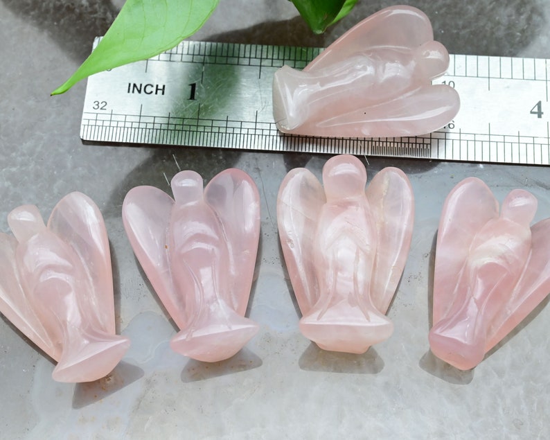 Puede incluir: Cuatro figuras de &aacute;ngeles de cuarzo rosa. Los &aacute;ngeles est&aacute;n tallados con alas detalladas y t&uacute;nicas fluidas. Una regla muestra que los &aacute;ngeles miden aproximadamente 2,5 cm de altura. Los &aacute;ngeles se exhiben sobre una superficie gris claro.