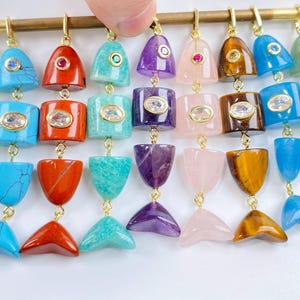 Può includere: Una collezione di nove ciondoli a forma di pesce appesi a una barra color oro. Ogni ciondolo è realizzato con pietre preziose di diversi colori, tra cui opale, turchese, diaspro rosso, amazzonite, ametista, quarzo rosa, occhio di tigre e giada.
