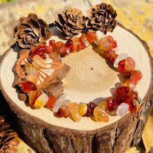 May include: A bracelet made of polished carnelian gemstones in shades of orange, red, and yellow. The bracelet is displayed on a wooden slice with three pinecones in the background. The gemstones are irregularly shaped and strung together.