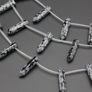 May include: A strand of black and white snowflake obsidian gemstone beads, each bead is a long, pointed, teardrop shape. The beads are strung on a clear elastic cord.