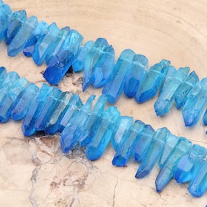 May include: A strand of blue crystal quartz points, also known as blue phantom quartz, arranged in a row on a light brown surface. The crystals are translucent and have a light blue color.