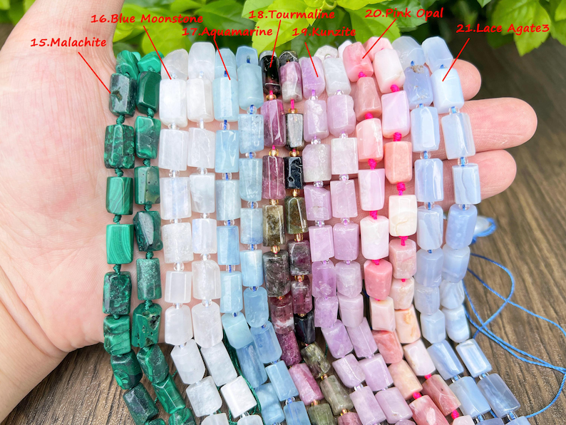 May include: A hand holds strands of gemstone beads in various colors. The beads are rectangular and strung together. Colors include green malachite, white, light blue aquamarine, pink, and lavender tourmaline, and pink opal. The beads are for jewelry making.