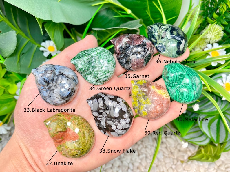 May include: A collection of ten small, polished, carved stone animal figurines. The figurines are in various colors and patterns, including green, red, black, white, and gray. The stones are labeled with their names, including Green Quartz, Black Labradorite, Snow Flake, Red Quartz, Malachite, Garnet, Nebula Stone, and Unakite.