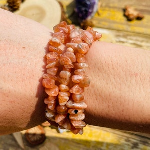 May include: A stack of three bracelets made of small, irregular, orange-brown sunstone beads. The bracelets are worn on a wrist, showcasing the natural variations in the stones. The beads have a slightly translucent quality, catching the light.