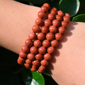 Puede incluir: Un conjunto de pulseras de cuentas de piedra arenisca roja. Las cuentas son redondas y tienen una superficie rugosa y texturizada.