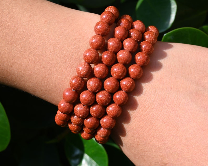 Puede incluir: Un conjunto de pulseras de cuentas de piedra arenisca roja. Las cuentas son redondas y tienen una superficie rugosa y texturizada.