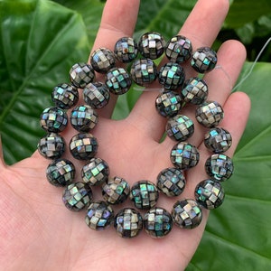 May include: A strand of round, iridescent, black and green shell beads with a checkered pattern. The beads are strung on a clear thread.