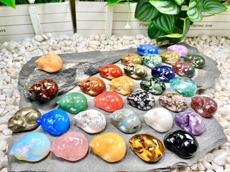 May include: A collection of 25 sleeping cat figurines carved from various types of polished stones, including amethyst, tiger's eye, rose quartz, and obsidian.