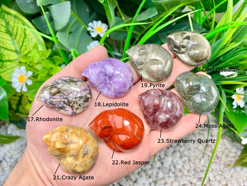 May include: A collection of 10 small, carved sleeping cat figurines made from various types of polished stones, including rhodonite, lepidolite, pyrite, gold obsidian, moss agate, strawberry quartz, crazy agate, and red jasper. Each figurine is labeled with its corresponding stone type.