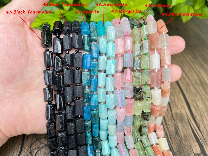 May include: A hand displays strands of colorful gemstone beads. The beads include black tourmaline, raw tourmaline, apatite, amazonite, morganite, prehnite, and mix moonstone. Each bead is a rectangular shape and strung together.