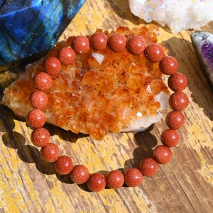 Puede incluir: Un brazalete de cuentas de piedra arenisca roja con una textura natural y rugosa. El brazalete est&aacute; sobre un grupo de cristales naranjas.