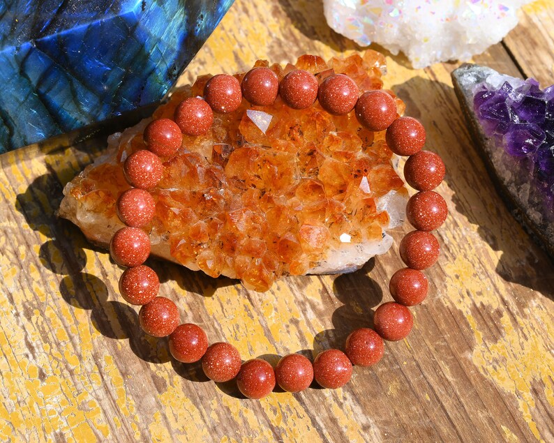 Puede incluir: Un brazalete de cuentas de piedra arenisca roja con una textura natural y rugosa. El brazalete est&aacute; sobre un grupo de cristales naranjas.