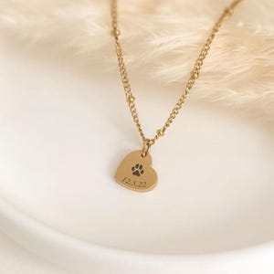 May include: A gold-toned necklace featuring a delicate chain with small beads and a heart-shaped pendant. The pendant is engraved with a black paw print graphic and the date 12.5.22, creating a simple and personalised piece of jewellery.