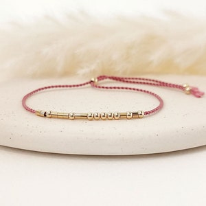 May include: A delicate bracelet with small, gold-coloured beads and cylindrical bars strung on a pink cord. The adjustable bracelet is displayed on a white, rounded surface, with a soft, blurred background.