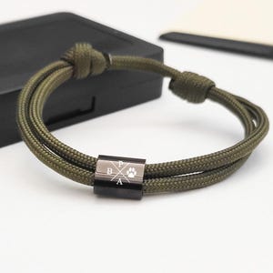 May include: An olive green rope bracelet with a black metal clasp. The clasp has the letters "B", "A", and "P" with a paw print symbol. The bracelet is knotted at both ends.
