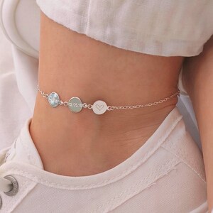 May include: A silver ankle bracelet with three round charms. The bracelet is on a person's ankle, and the charms are visible.