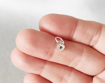 May include: A small, silver-colored heart charm with the letter "P" engraved on it. The charm is attached to a small ring, designed to be added to a necklace or bracelet. The charm is held in a person's hand.