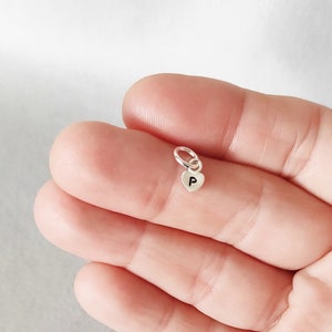 May include: A small, silver heart charm with the letter "P" engraved on it. The charm is attached to a small ring, designed to be added to a necklace or bracelet. The charm is held in a person's hand.