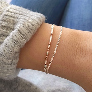 Könnte beinhalten: Ein zartes silbernes Kettenarmband, das mit einem Perlenarmband mit abwechselnden silbernen und kupfernen Perlen übereinander gelegt wird.