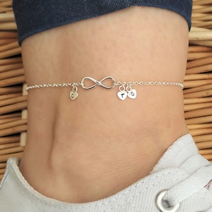 May include: Silver infinity anklet with small heart charms, each with a letter. The anklet is on a person's ankle, with a white trainer.