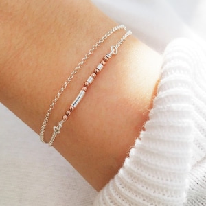 May include: A silver chain bracelet with a rose gold bar and beads. The bracelet is layered on top of another silver chain bracelet.